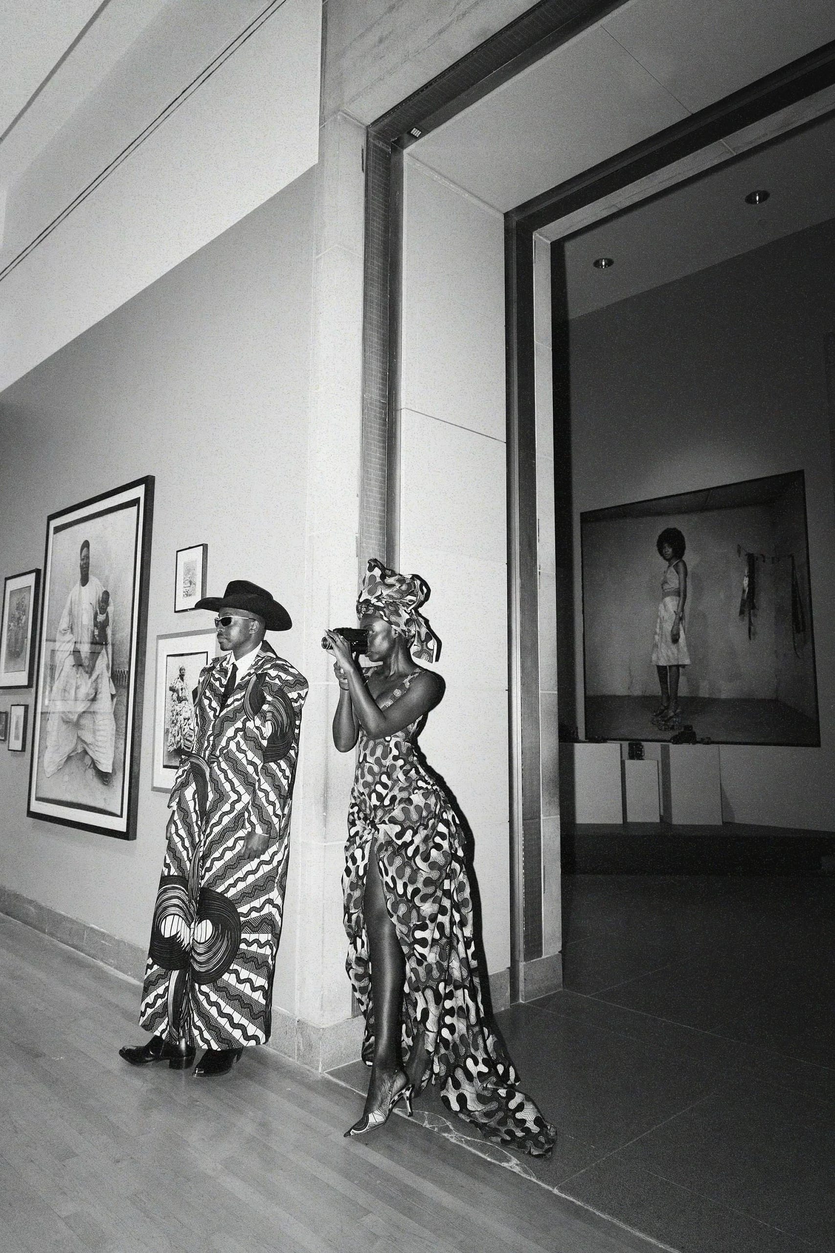 A black-and-white portrait of Trevor and Diouf standing side by side, with Diouf taking a photo using her camera.