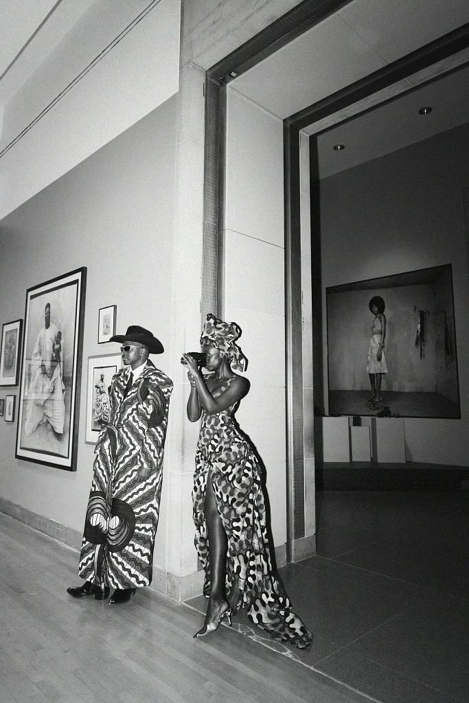 A black-and-white portrait of Trevor and Diouf standing side by side, with Diouf taking a photo using her camera.